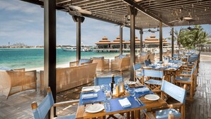 A seaside restaurant with wooden tables and chairs, a view of the ocean, and a building with a red roof in the background. - Anantara Palm Jumeirah Luxury 1BR Sea View (Dubai)