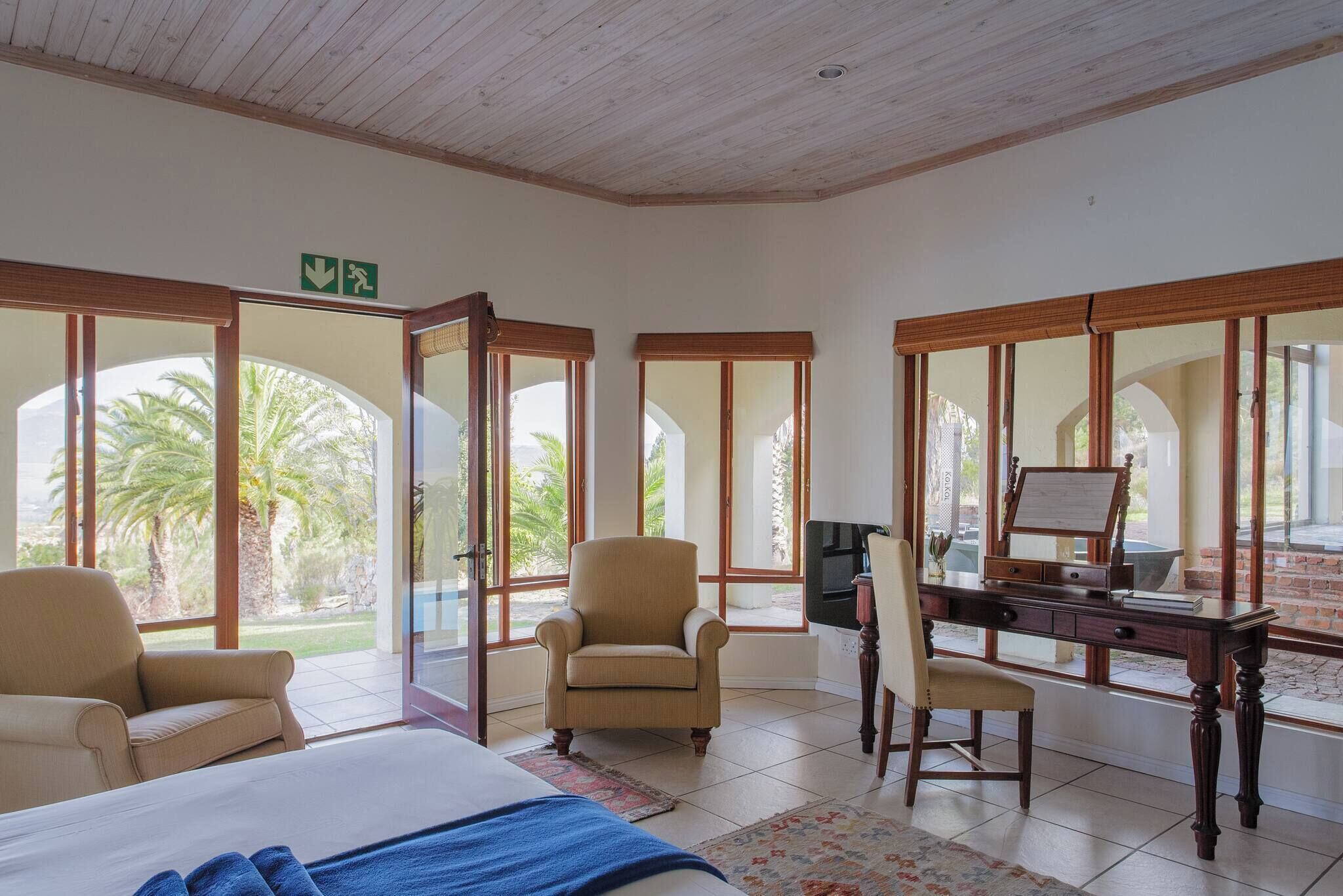 Camera d'albergo con un letto, una scrivania, una sedia e ampie finestre con vista su palme.