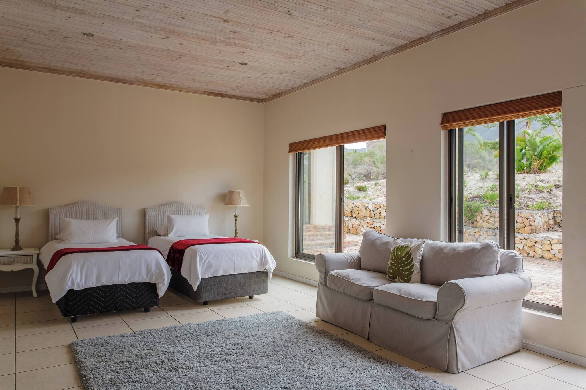 A bedroom with two beds, a grey sofa, and large windows with a view of a garden.