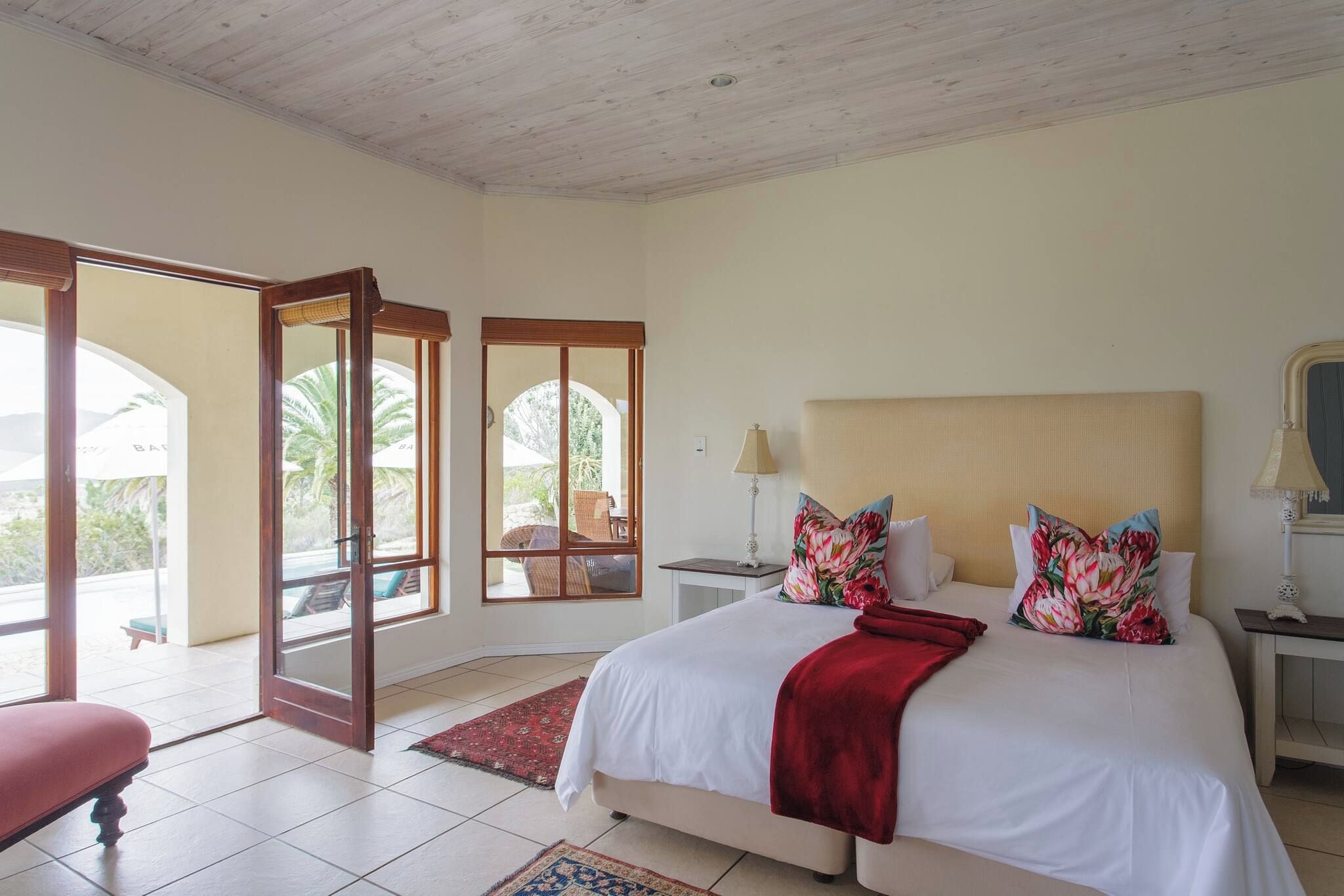 A bedroom with a large bed, a red bench, a nightstand with a lamp, and a view through a glass door to an outdoor area.