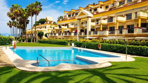 A swimming pool surrounded by a well-maintained lawn and palm trees, with a multi-story building in the background. - Apartment 'En Elviria' with Shared Pool, Wi-Fi and Air Conditioning (Marbella)