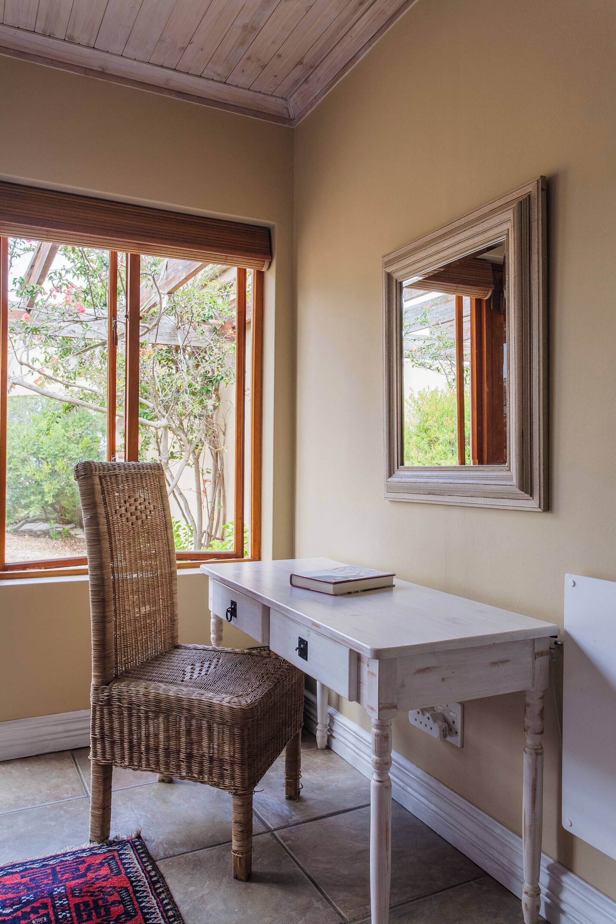 A room with a wicker chair, a white desk, a mirror, and a window with a view of greenery.