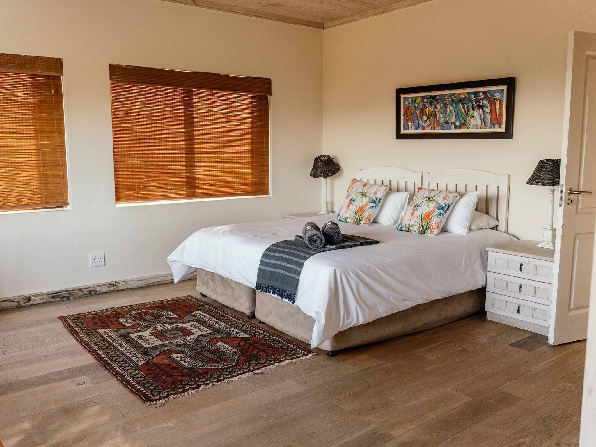 Une chambre à coucher avec un lit, des tables de chevet, un tapis, un tableau et une fenêtre avec des stores.