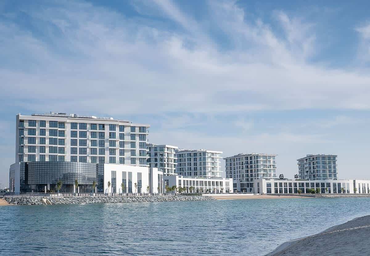 Un hôtel situé au bord de l’eau, composé de plusieurs immeubles de grande hauteur, avec une plage en arrière-plan et un ciel dégagé.
