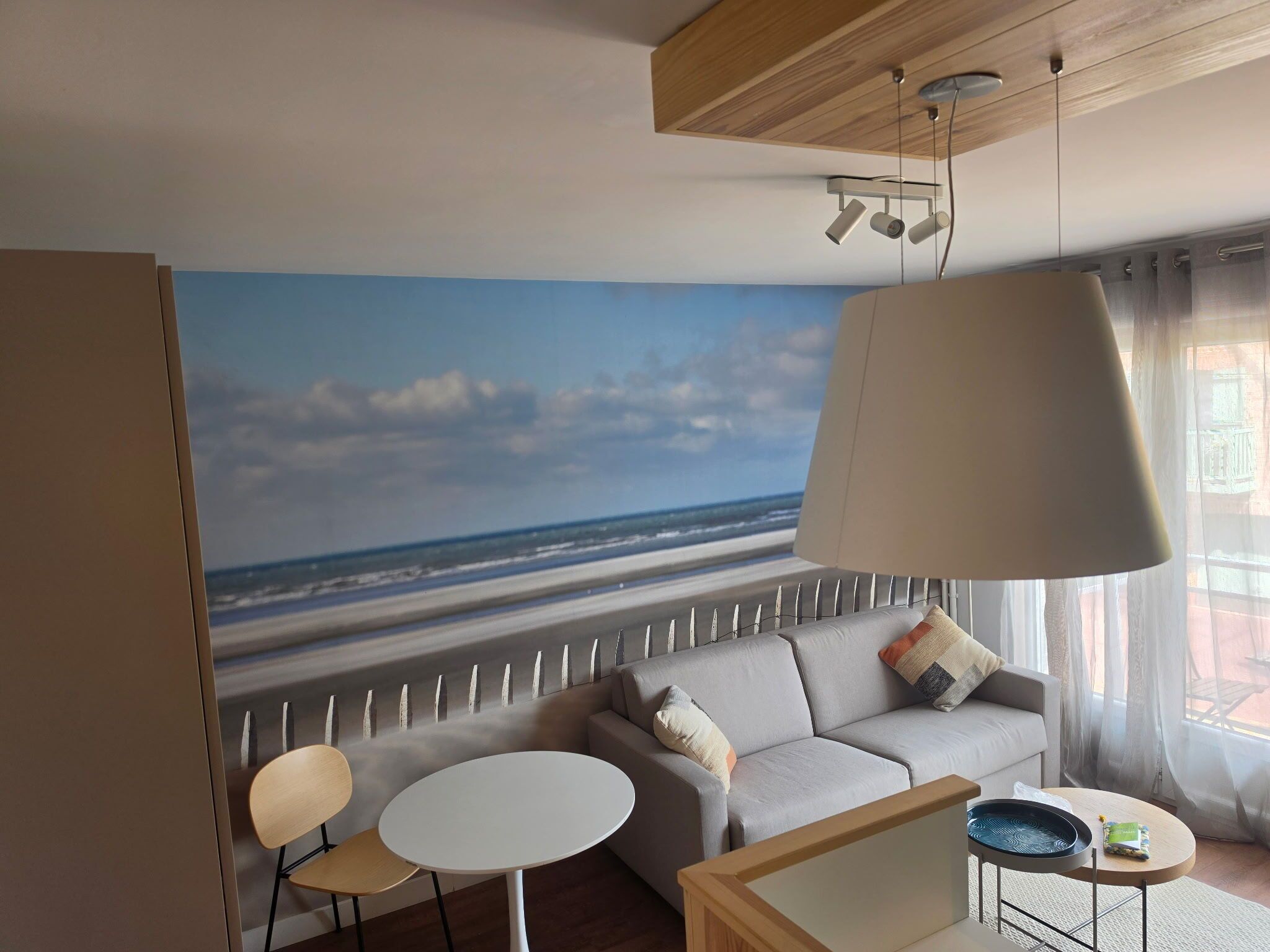 Uma sala de estar moderna com um grande mural de praia, um sofá, duas poltronas, uma mesa de centro e um abajur pendente.