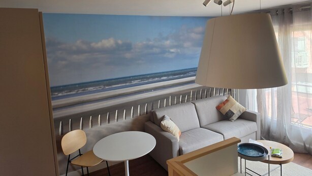 A modern living room with a large beach mural, a sofa, two chairs, a coffee table, and a hanging lamp. - Modern and functional apartment near the beach and downtown (Paris)