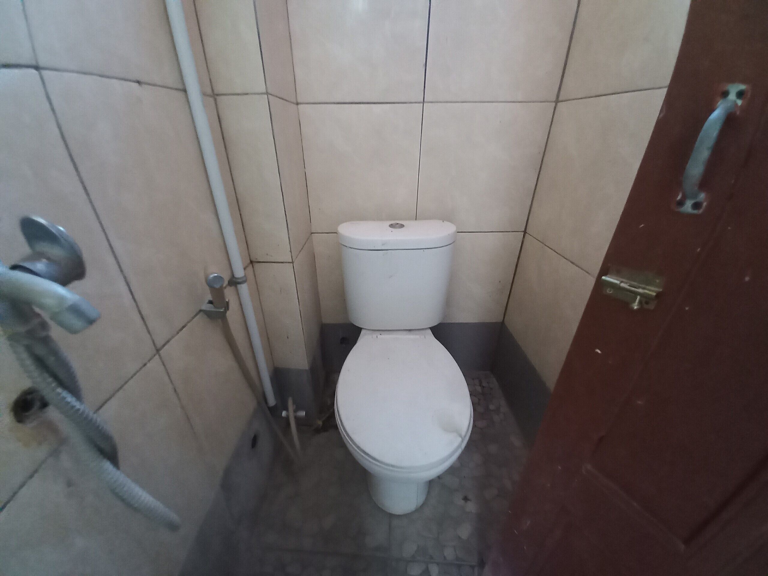Une petite salle de bain carrelée, avec des toilettes blanches, un tuyau en métal et une porte rouge.