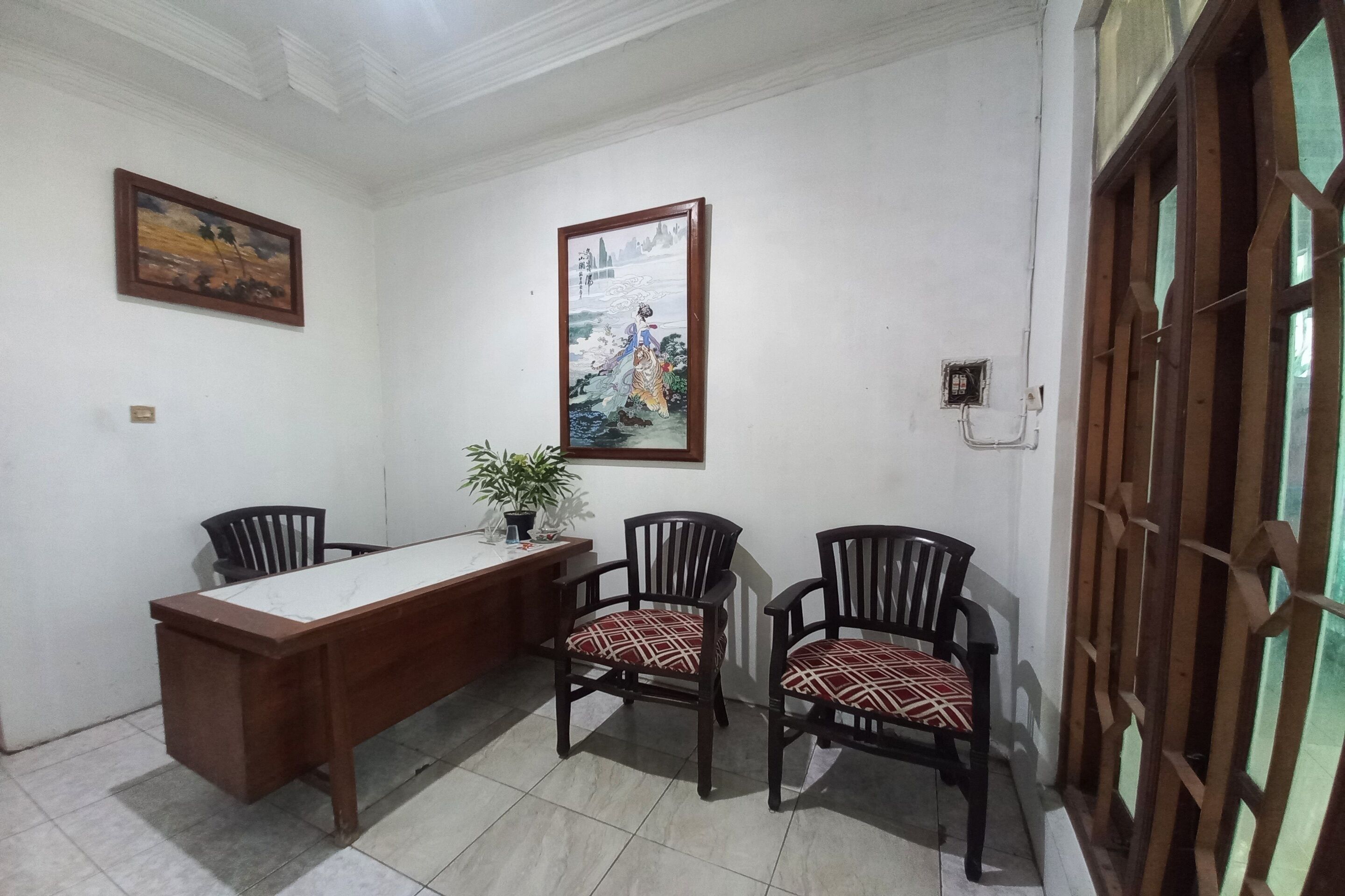 A room with a wooden desk, two chairs, a potted plant, and two framed artworks on the wall.