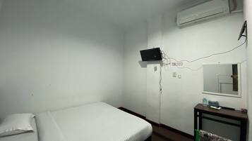 Una habitación pequeña y poco amoblada con una cama individual, un escritorio con una silla, aire acondicionado instalado en la pared y un televisor.
