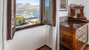 A room with a large window offering a view of a lake and mountains, a wooden dresser, and a framed picture on the wall. - Charming Bellagio Manzoni in the heart of Bellagio with beautiful lake view (Bellagio)