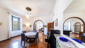 A spacious room with a wooden floor, a dining table with chairs, a piano, and a large window with curtains. - Charming Bellagio Manzoni in the heart of Bellagio with beautiful lake view (Bellagio)