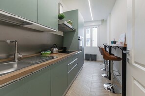 Eine moderne Küche mit Spüle aus Edelstahl, grüne Schränke und Dunstabzugshaube aus Edelstahl. An der Wand stehen Bartresen, und ein Fenster sorgt für natürliches Licht.