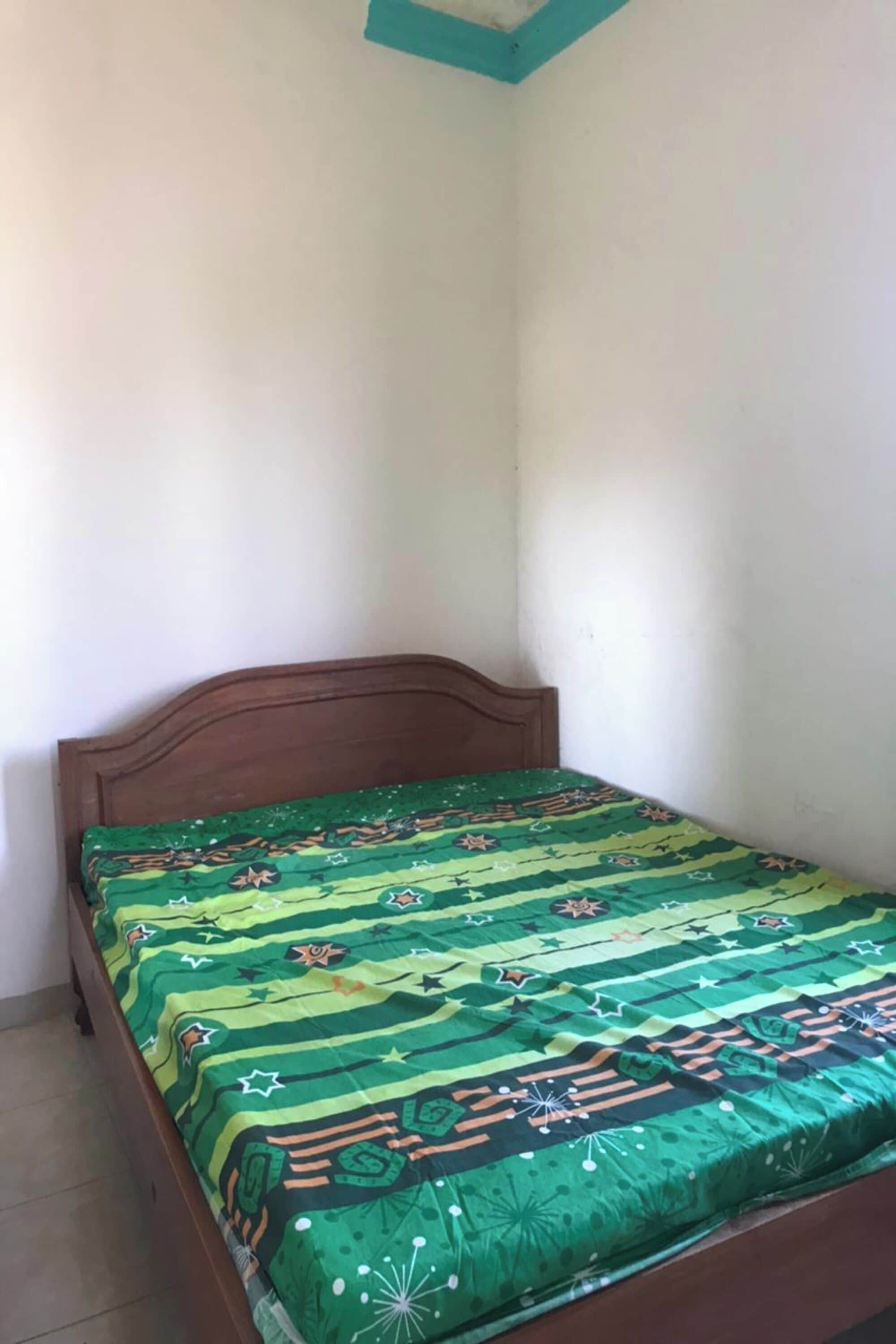 Una stanza piccola con un letto singolo, una testiera in legno, una coperta a letto con motivo verde e pareti bianche.