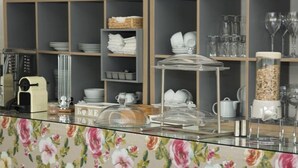 Ein Frühstücksbuffet mit floralem Muster, einer Kaffeemaschine sowie verschiedenen Gläsern und Geschirr.