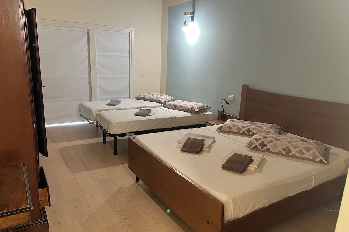 Habitación de hotel con dos camas individuales, una cama más grande, un armario de madera, un escritorio y una puerta que da a un balcón.