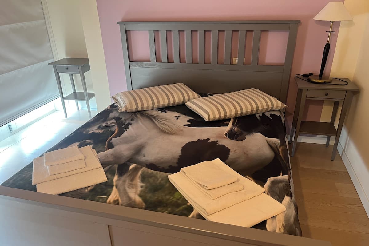 Una cama con una colcha estampada de caballos, dos almohadas, una lámpara de noche y una mesita.