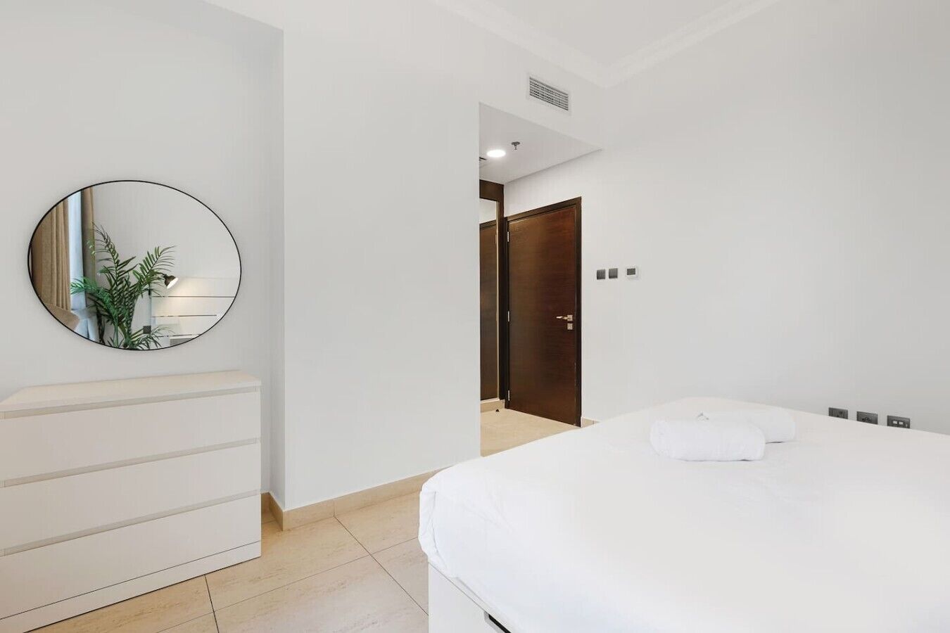 Une chambre d’hôtel avec un lit, un miroir rond, une commode blanche et une porte donnant sur une autre pièce.