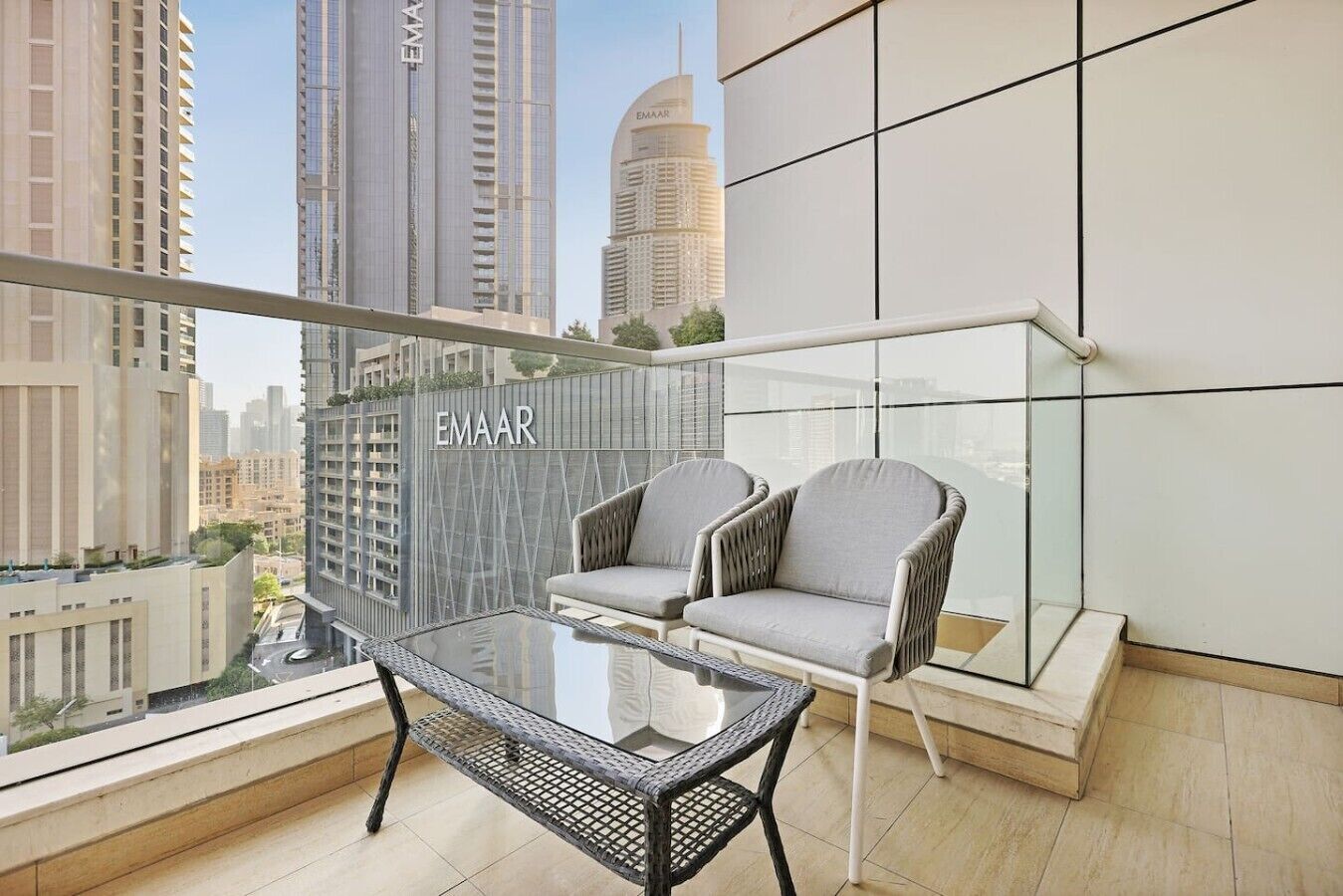 A balcony with a glass table and two chairs, overlooking a cityscape with high-rise buildings.
