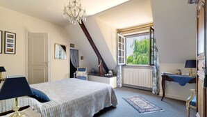 A bedroom with a bed, a desk, a chair, a chandelier, a window, and a radiator.
