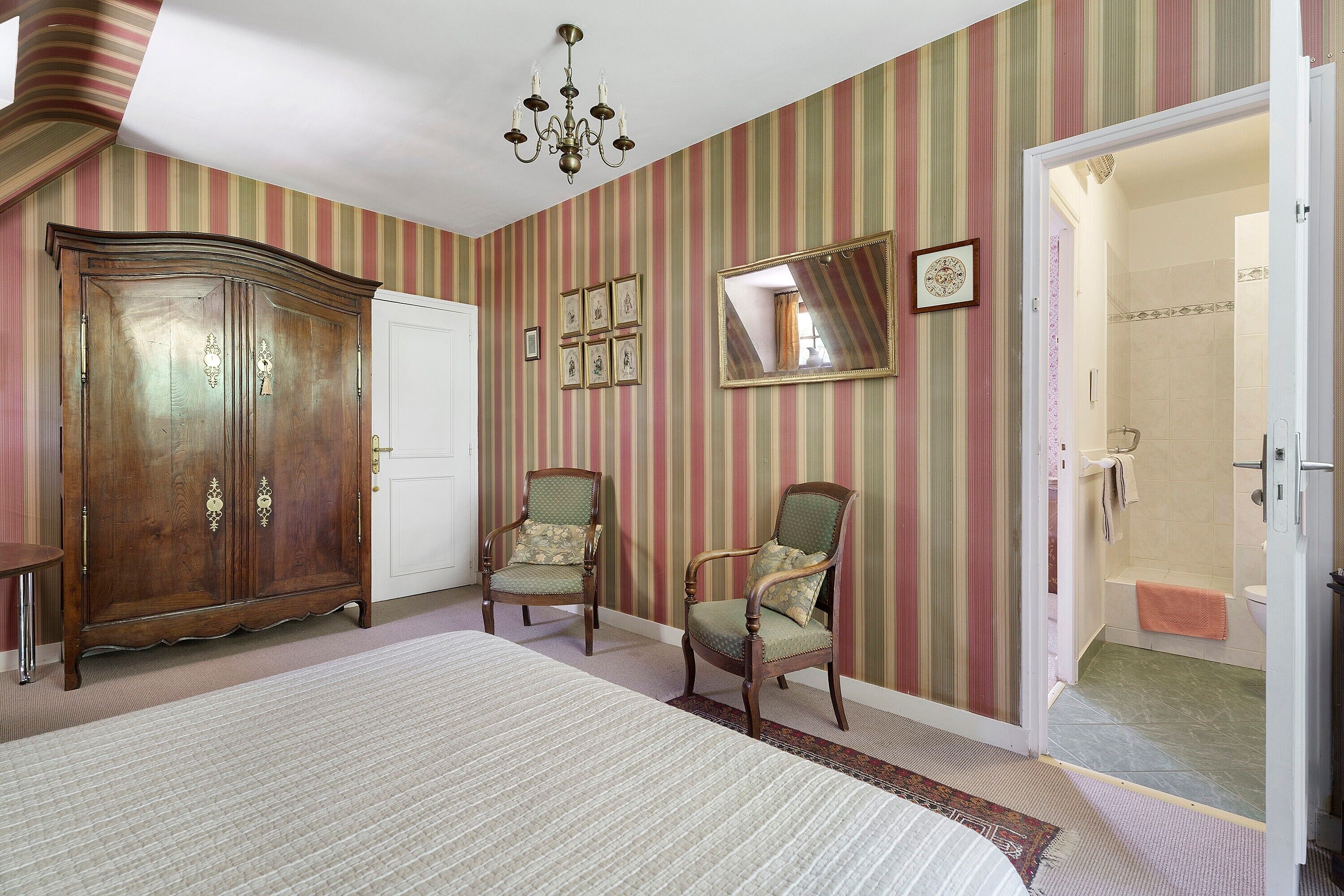 A room with a large wooden wardrobe, two upholstered chairs, a mirror, and a chandelier.