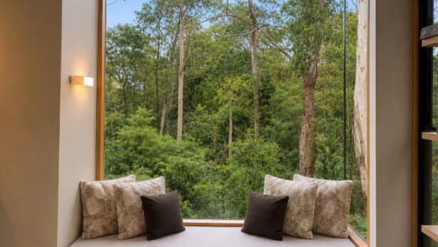 Ein Zimmer mit großem Fenster und Blick auf einen Wald. Es gibt ein Bett mit zwei Kissen und eine Wandleuchte.
