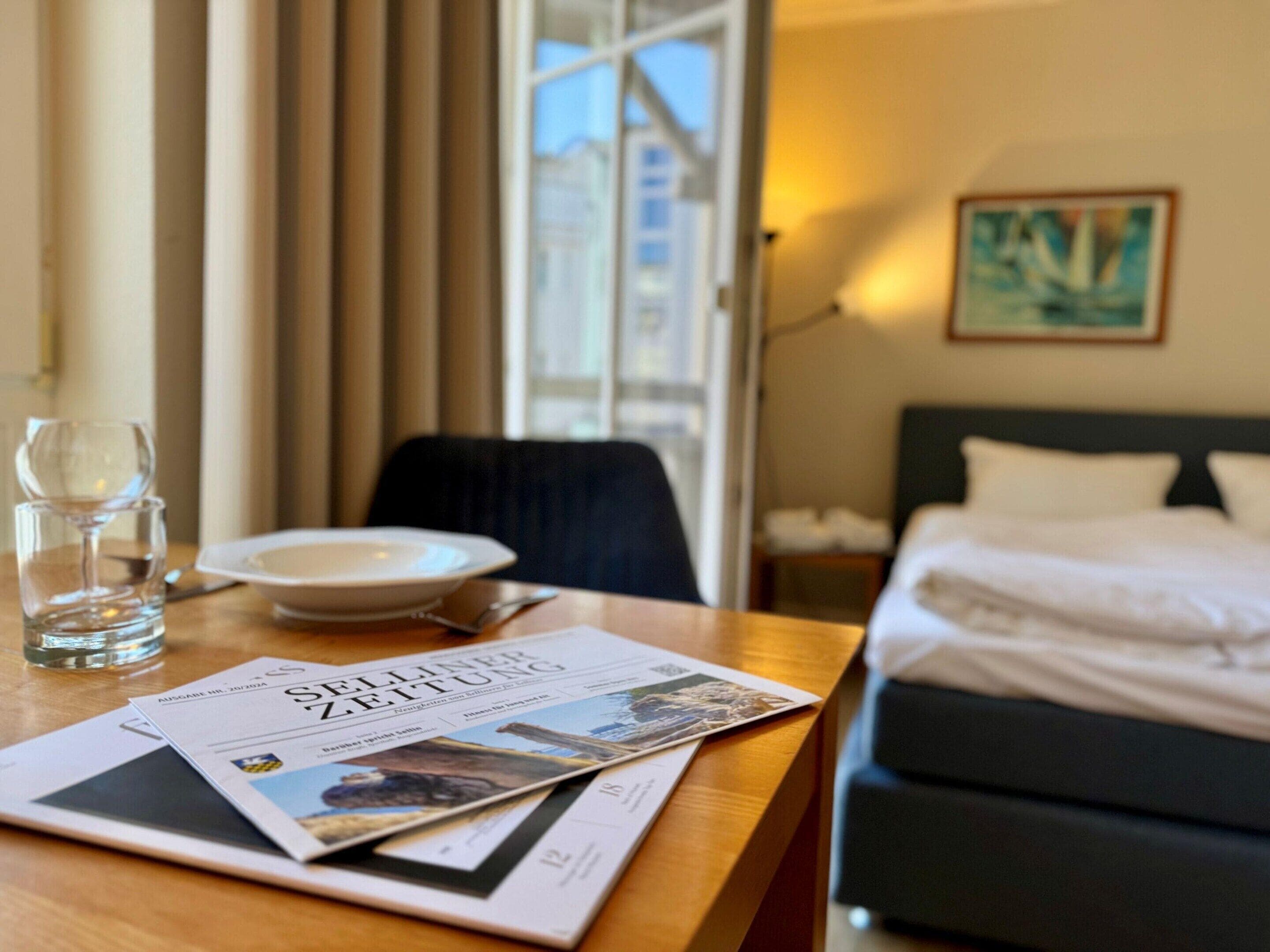 Habitación de hotel con cama, una mesa de comedor, un vaso de agua, un plato y un periódico.