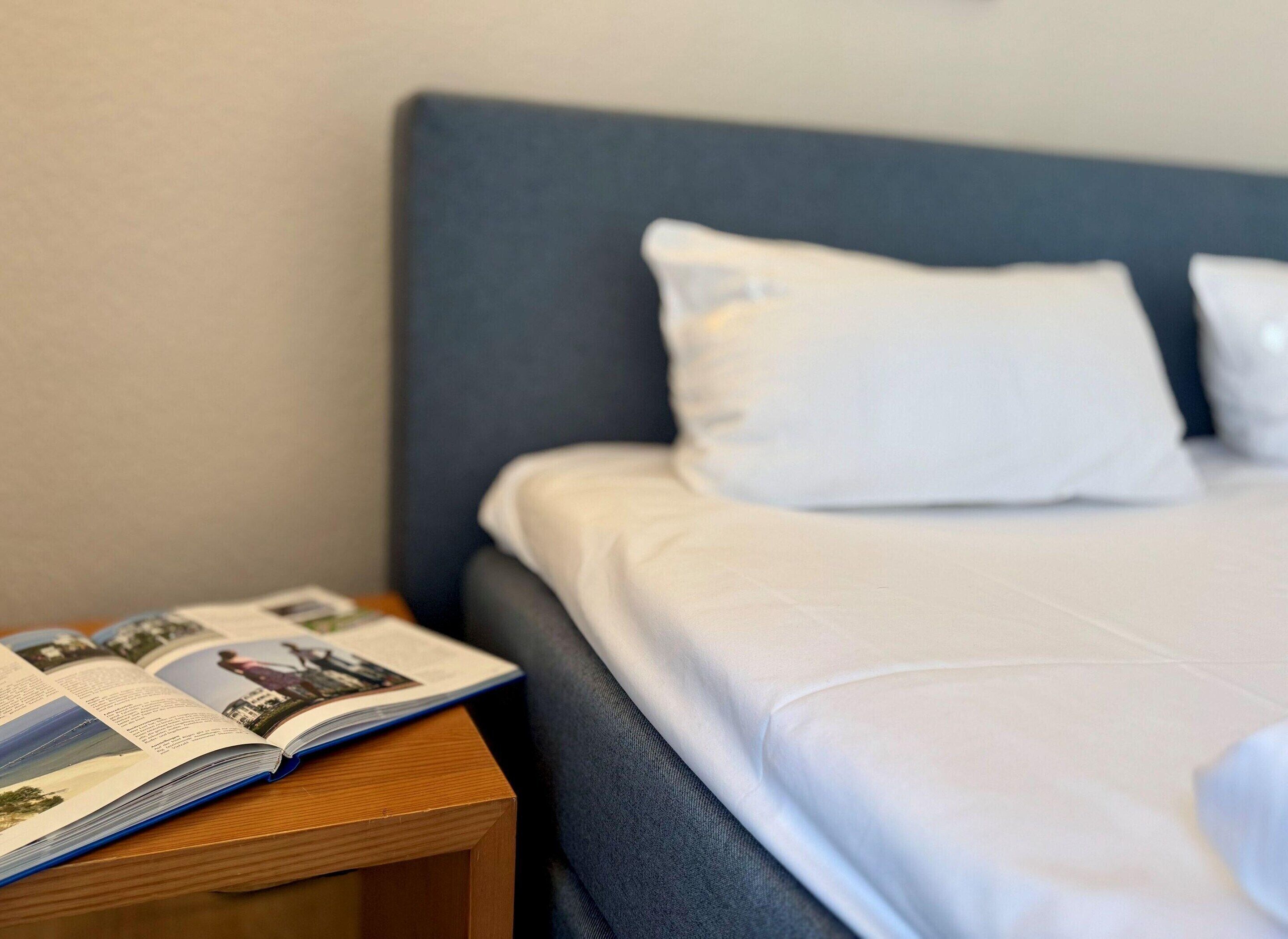 Ein ordentlich bezogenes Bett mit weißer Bettwäsche und Kissen, ein Nachttisch aus Holz mit einem offenen Buch und ein dunkelblaues Kopfstück.