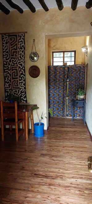 A room with a wooden floor, a dining table with chairs, a blue potted plant, a patterned curtain, and a wall-mounted light fixture.