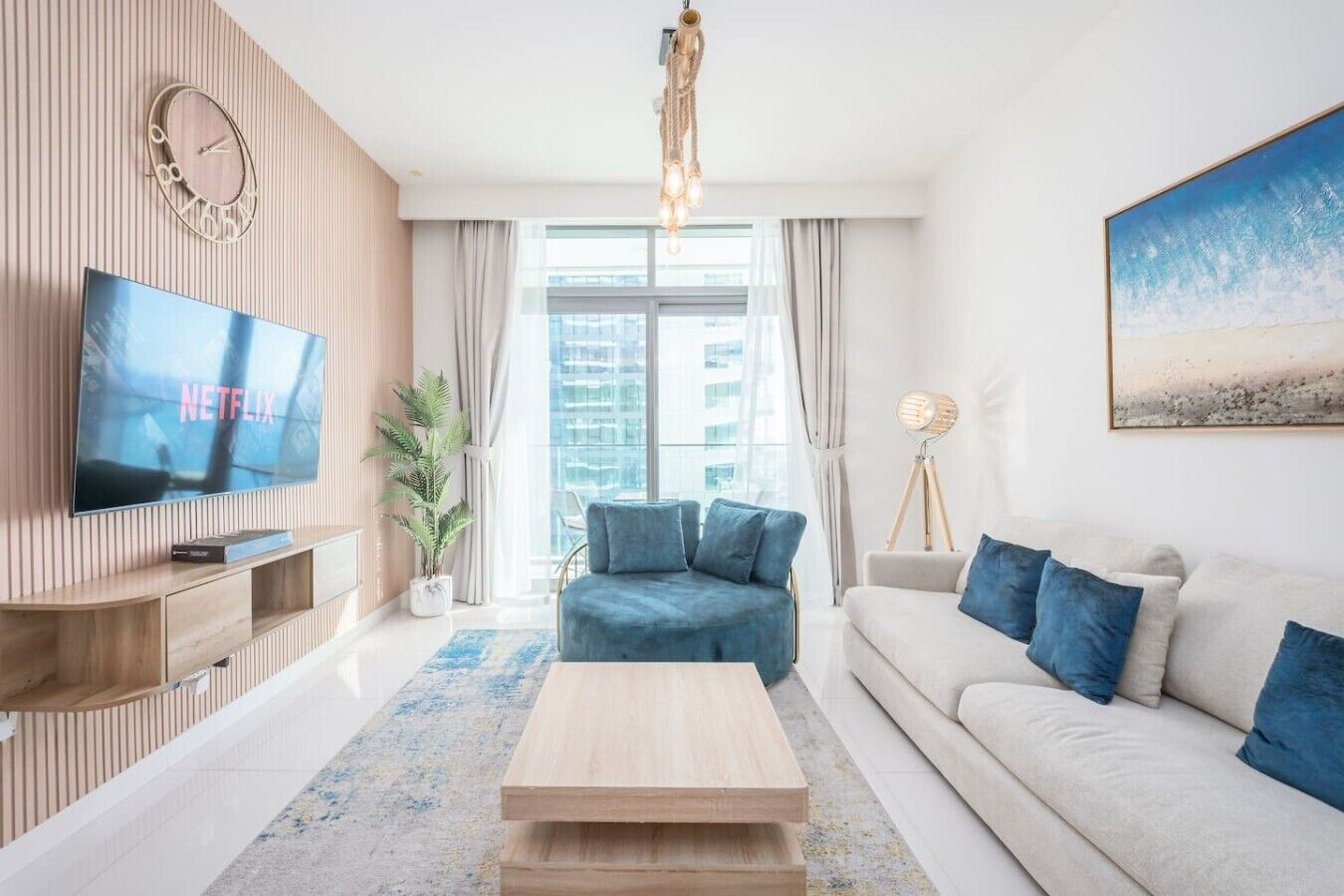 Een moderne woonkamer met een flatscreen televisie, een houten tv-meubel, een blauwe bank, een witte bank, een houten salontafel, een vloerlamp en een groot raam met gordijnen.