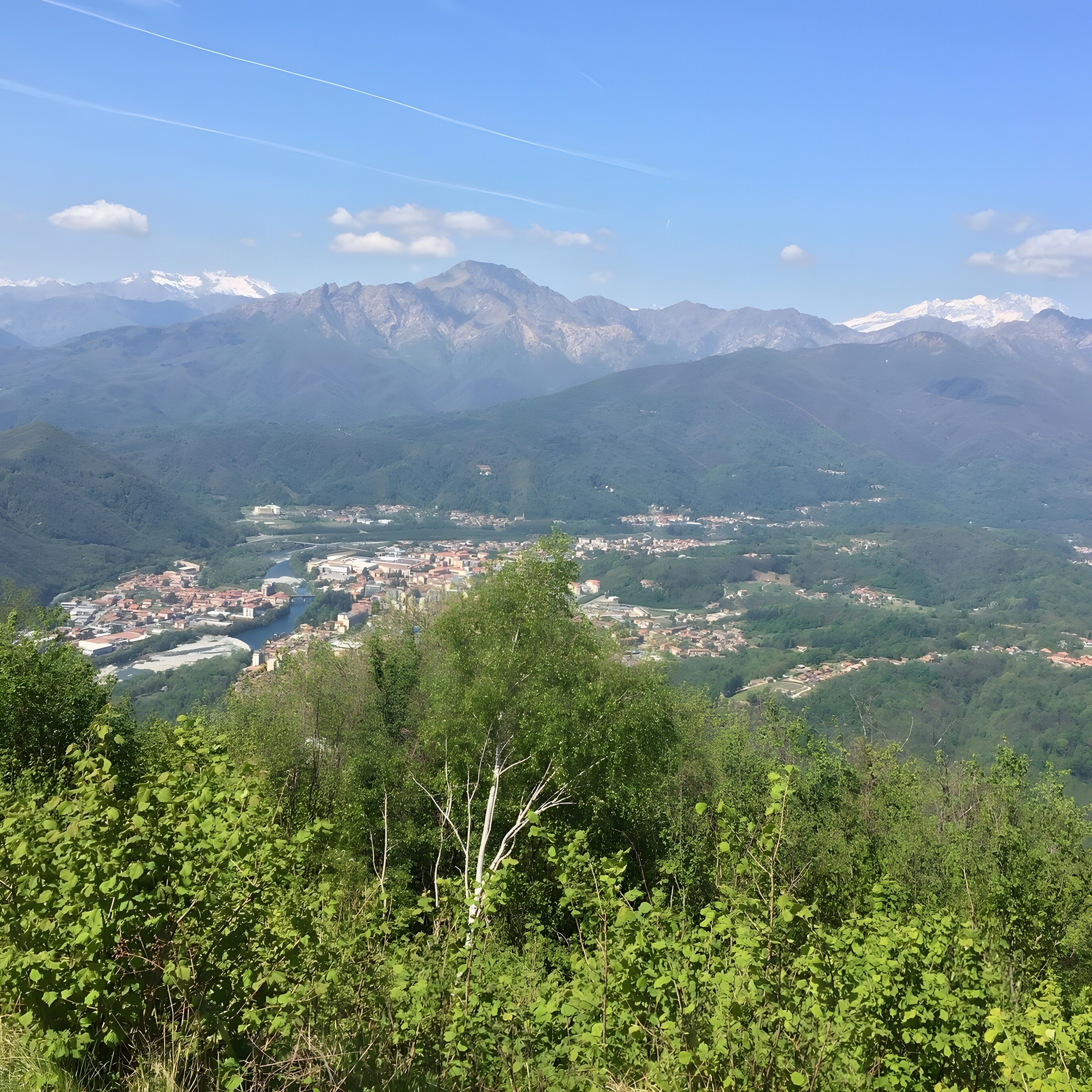 Eine gebirgige Landschaft mit einer Stadt, eingebettet in ein Tal, umgeben von viel Grün und fernen, schneebedeckten Gipfeln.