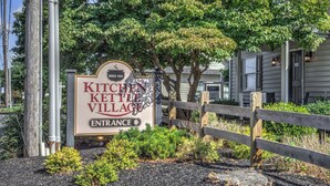 Un cartel que dice 'KITCHEN KETTLE VILLAGE' con una flecha indicando la entrada, rodeado de árboles y una valla de madera.