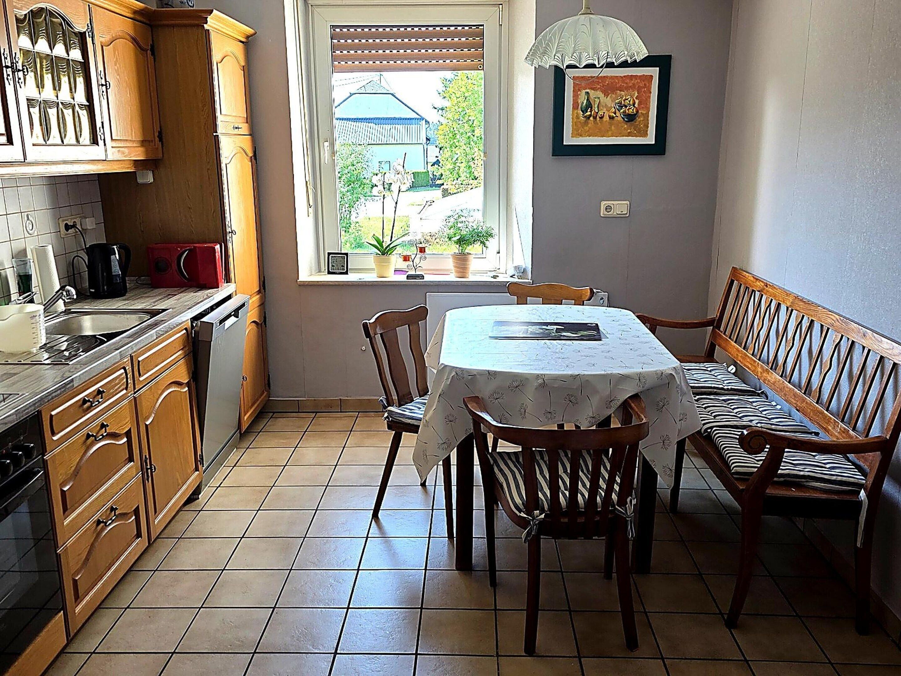 Une cuisine avec des meubles en bois, une table à manger avec des chaises, une fenêtre donnant sur un espace verdoyant et un luminaire suspendu.