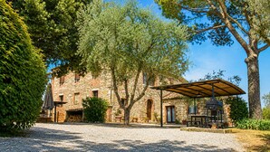 A stone building with a gravel driveway, a covered outdoor seating area, and a large tree. - Stunning "La Quercia" Apartment in a Country House with a Pool and Panoramic View (Toscana)