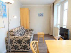 Ein Wohnzimmer mit einer Couch, einem kleinen Tisch, einem Sessel, einer Stehlampe, einem Fernseher und einem Fenster mit Vorhängen.