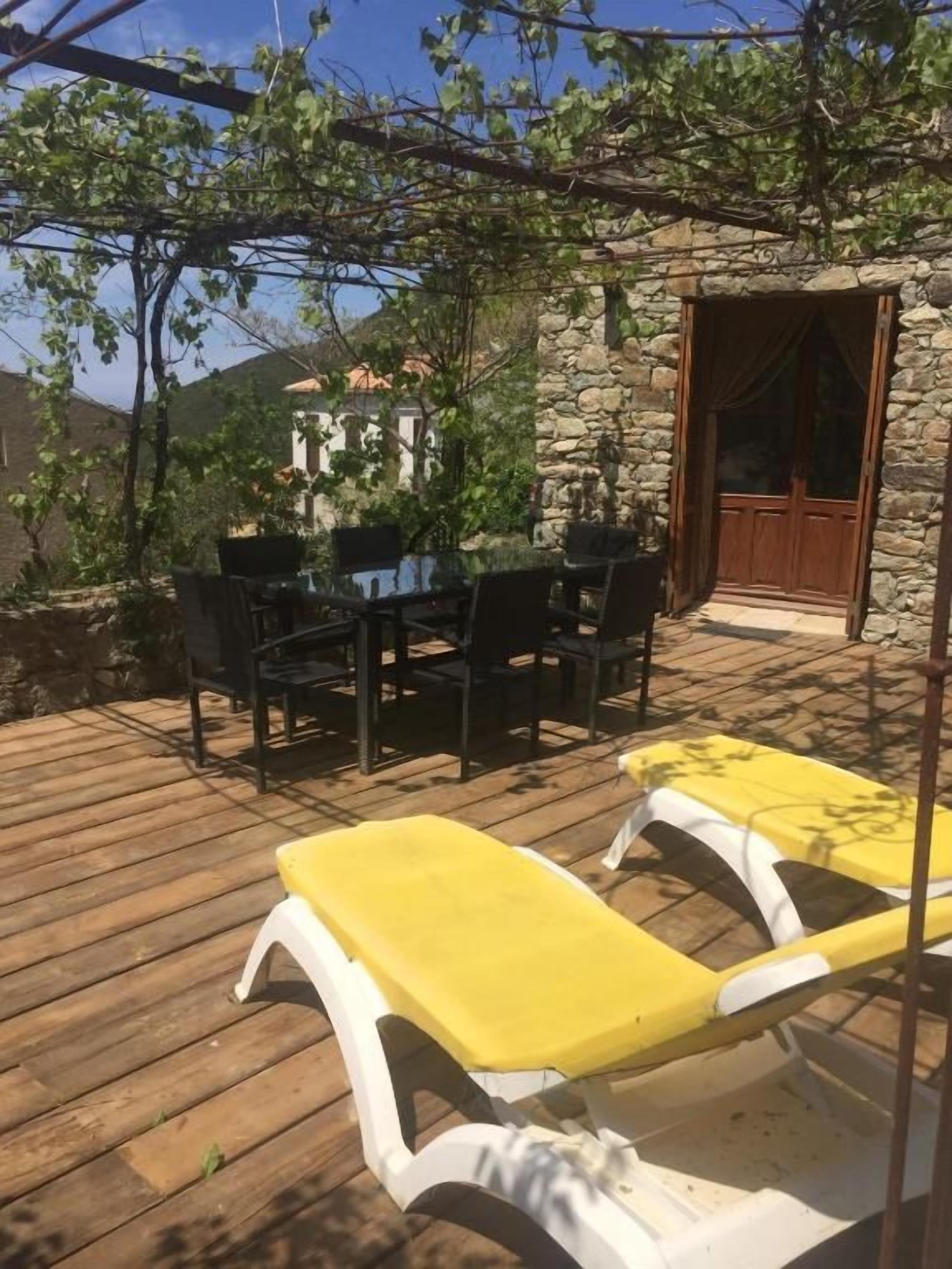 Une terrasse en bois avec une table et des chaises, un fauteuil de jardin jaune et un bâtiment en pierre avec une porte.