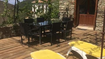 Um deck de madeira com uma mesa e cadeiras, uma poltrona amarela e um edifício de pedra com uma porta.