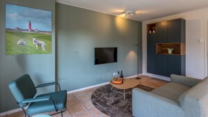 A living room with a flat-screen TV, a green sofa, a blue armchair, a round coffee table, and a painting of a lighthouse and sheep. - 4-guest Bungalow Near Texel Dunes (De Koog)
