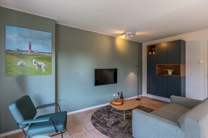 Ein Wohnzimmer mit einem Flachbildfernseher, einem grünen Sofa, einem blauen Sessel, einem runden Couchtisch und einem Bild von einem Leuchtturm und Schafen.