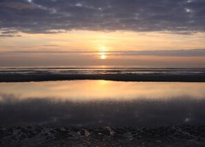 Ein Strandbild mit untergehendem Sonnenuntergang am Horizont, ruhigem Wasser, das den Himmel spiegelt, und freier Sicht auf den Strand.