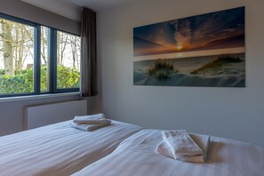 Ein Hotelzimmer mit großem Fenster, einem Bett mit weißer Bettwäsche und einem Sonnenuntergangsgemälde an der Wand.