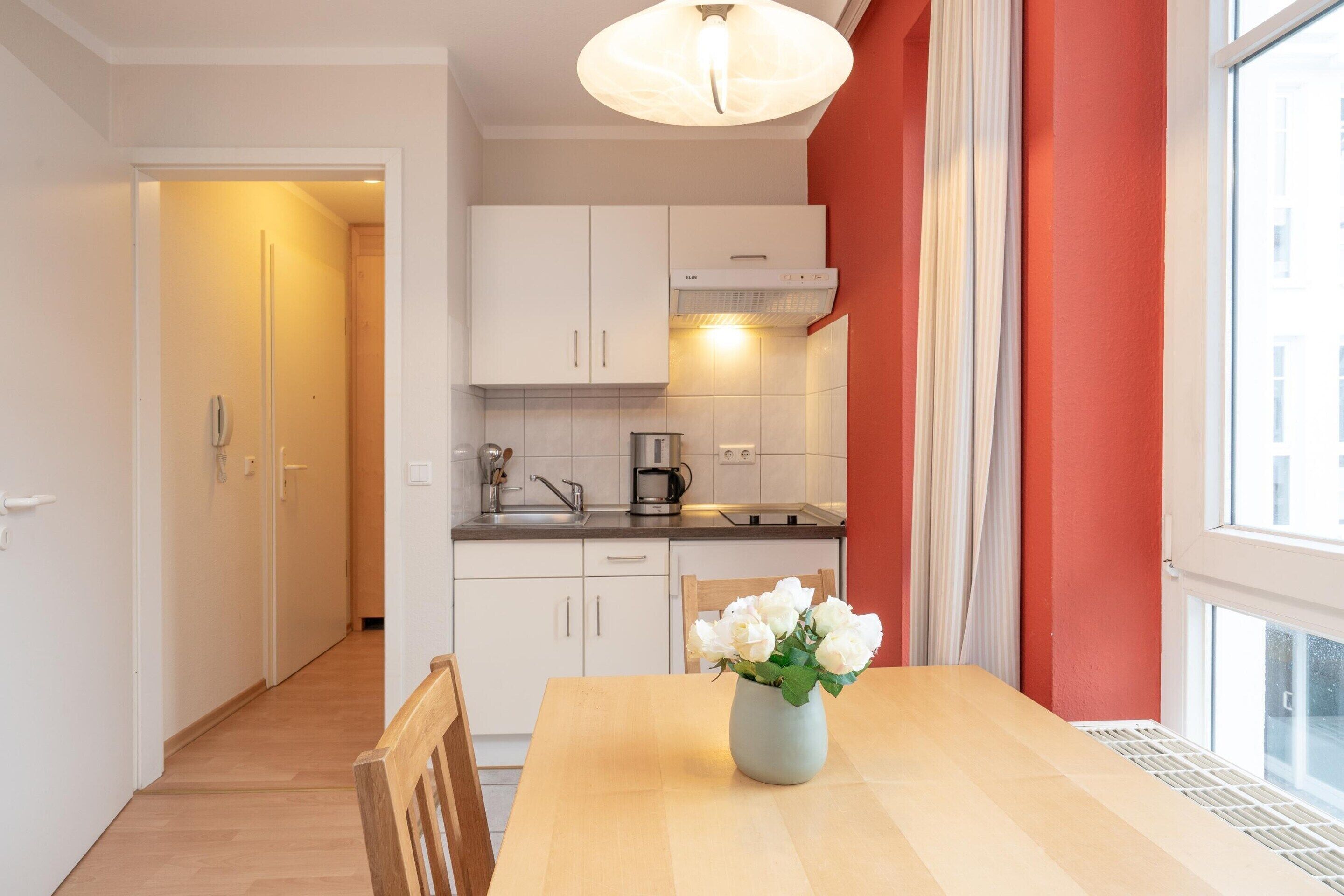 A kitchen with white cabinets, a wooden table with a vase of flowers, and a window with curtains.