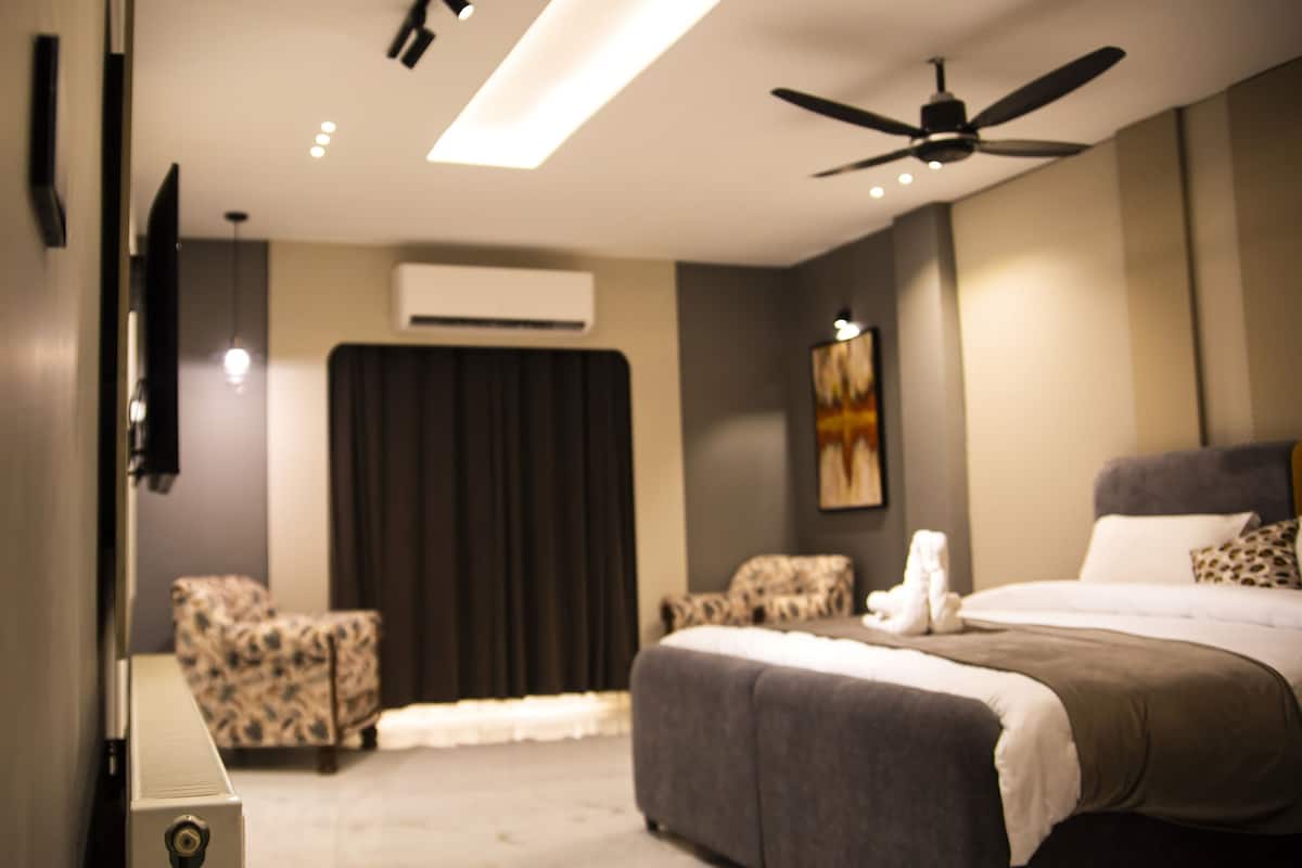 A hotel room with a bed, two chairs, a ceiling fan, a flat-screen TV, and an air conditioning unit.