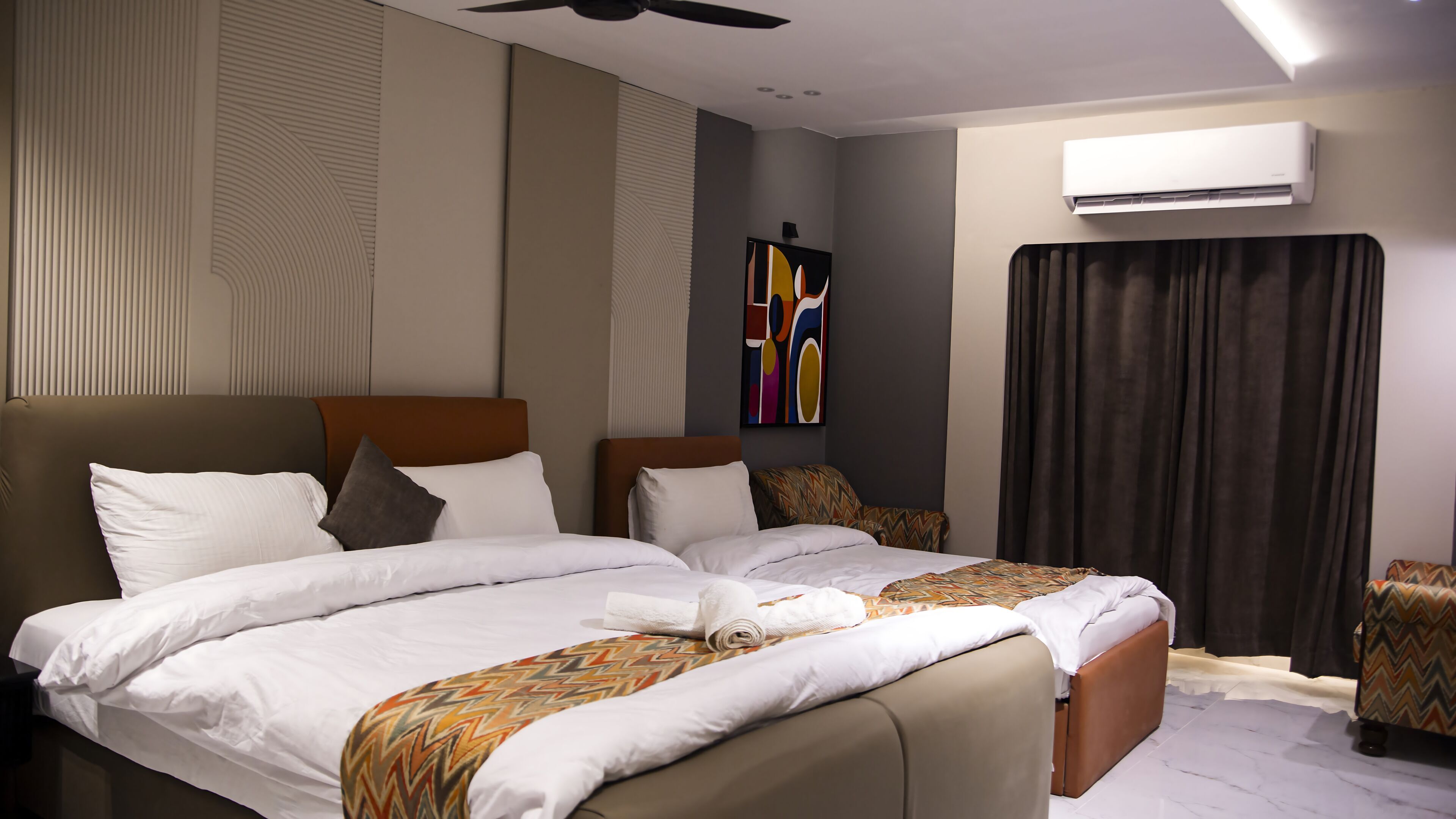 Quarto de hotel com uma cama grande, ventoinha de teto, ar condicionado e uma pintura abstrata moderna.