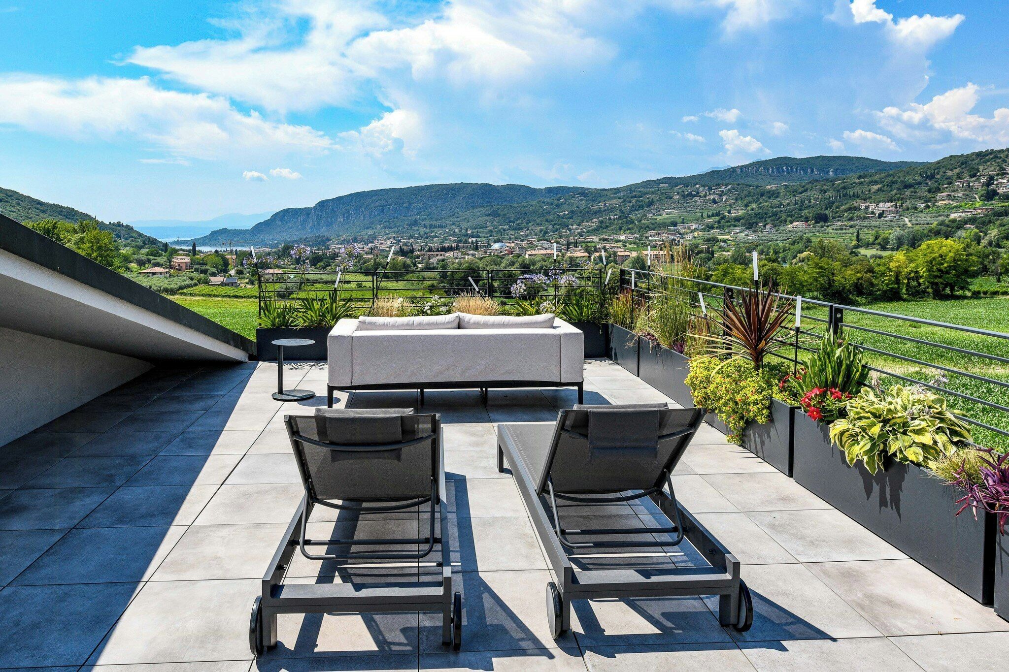 Eine Dachterrasse mit einer Couch, zwei Liegestühlen und Blick auf ein grünes Tal und Berge.