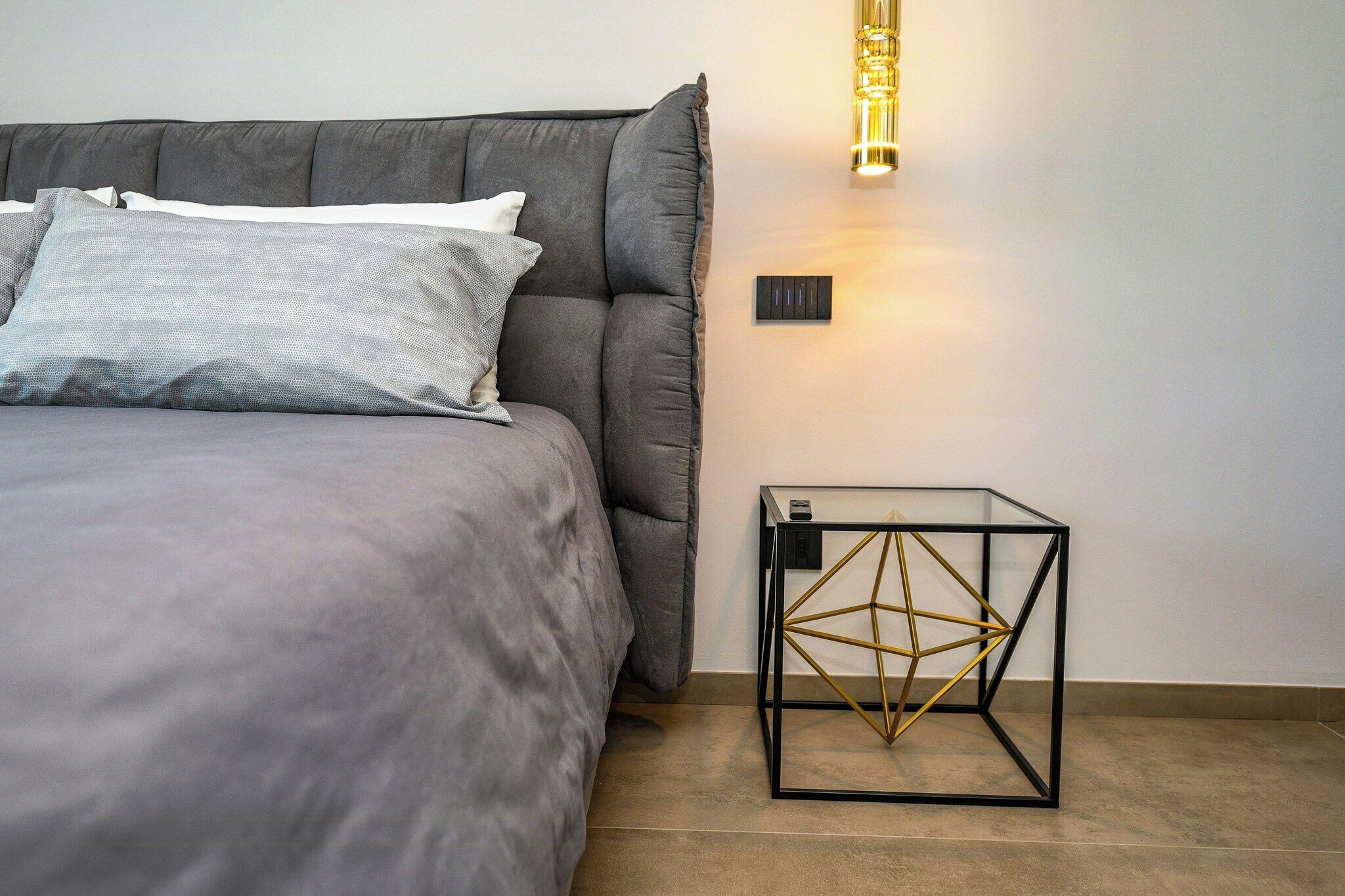 A modern bedroom with a bed, pillows, a bedside table with a geometric design, and a wall-mounted light fixture.
