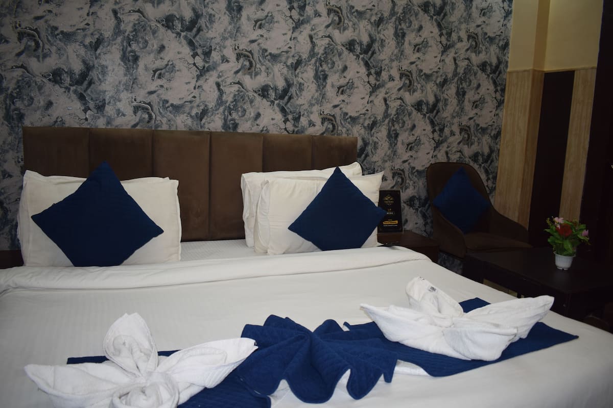 A hotel room with a bed, pillows, a nightstand, a chair, and a potted plant.