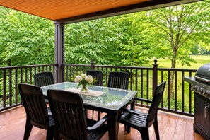 Eine überdachte Terrasse im Freien mit einem Glastisch, schwarzen Stühlen, einem Grill und Blick auf eine grüne Landschaft.