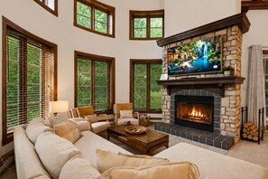 A living room with a stone fireplace, large windows, and a comfortable seating area. - Tremblant Prestige - Manoir 112-10 (Mont-Tremblant)