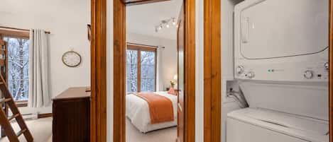 A bedroom with a bed, a wooden dresser, a washing machine, and a window with a view of snow-covered trees.