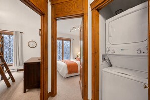 Ein Schlafzimmer mit einem Bett, einer Holzkommode, einer Waschmaschine und einem Fenster mit Blick auf schneebedeckte Bäume.